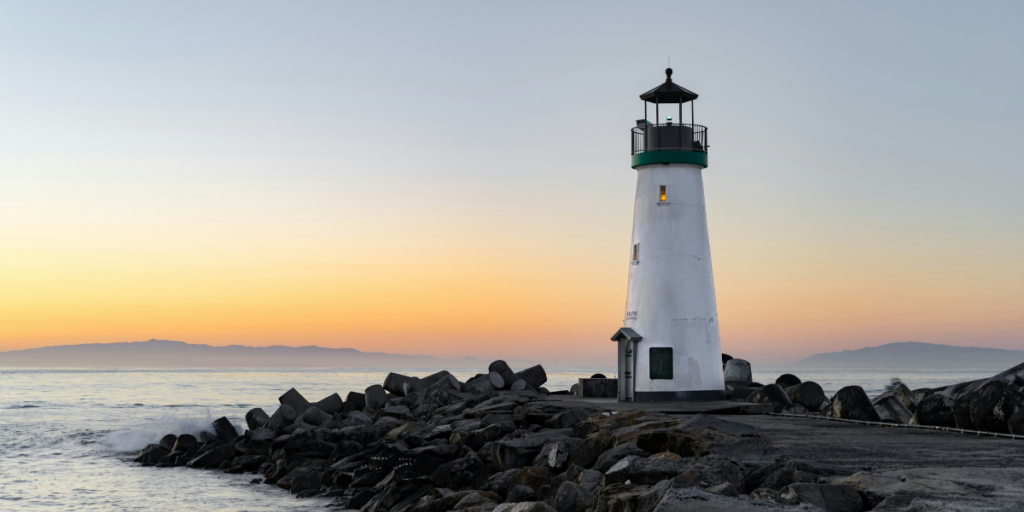 A lighthouse on the edge of an ocean with a view of a sunset.